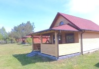 Drewniany domek letniskowy z czerwonym dachem i zadaszonym tarasem, otoczony zieloną trawą i drzewami